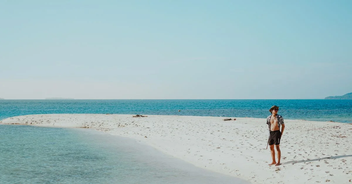 Un homme seul se tient détendu sur une plage de sable blanc pittoresque au bord d'eaux bleues claires, sous un ciel lumineux.