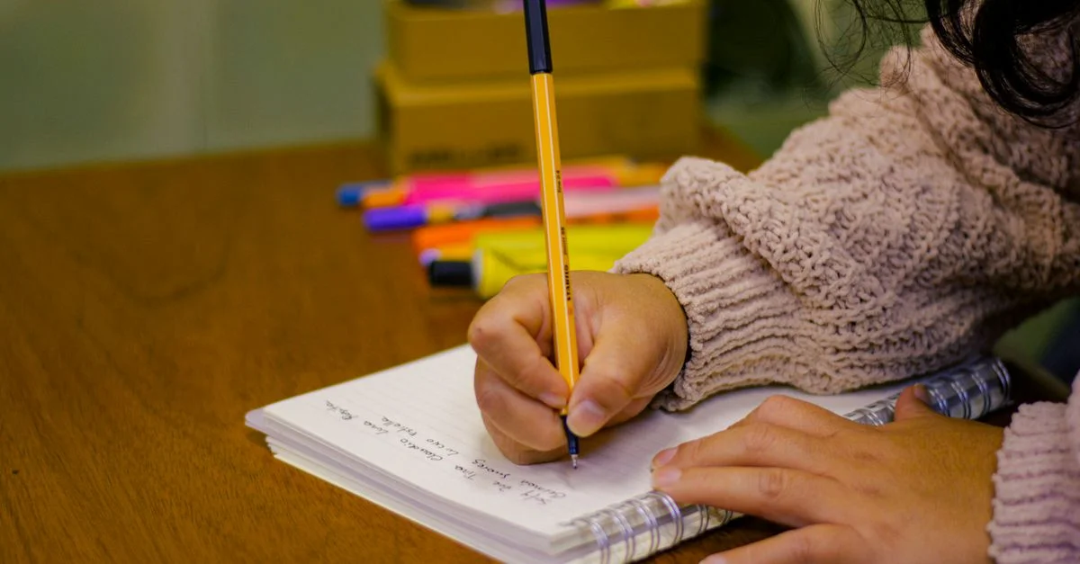 Une personne écrivant dans un cahier avec des stylos et des surligneurs sur un bureau en bois, créant une scène d'étude.